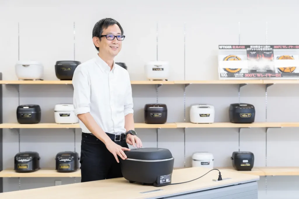 Mr. Meshizuka standing in front of a rice cooker (JRT-A) and speaking while touching it